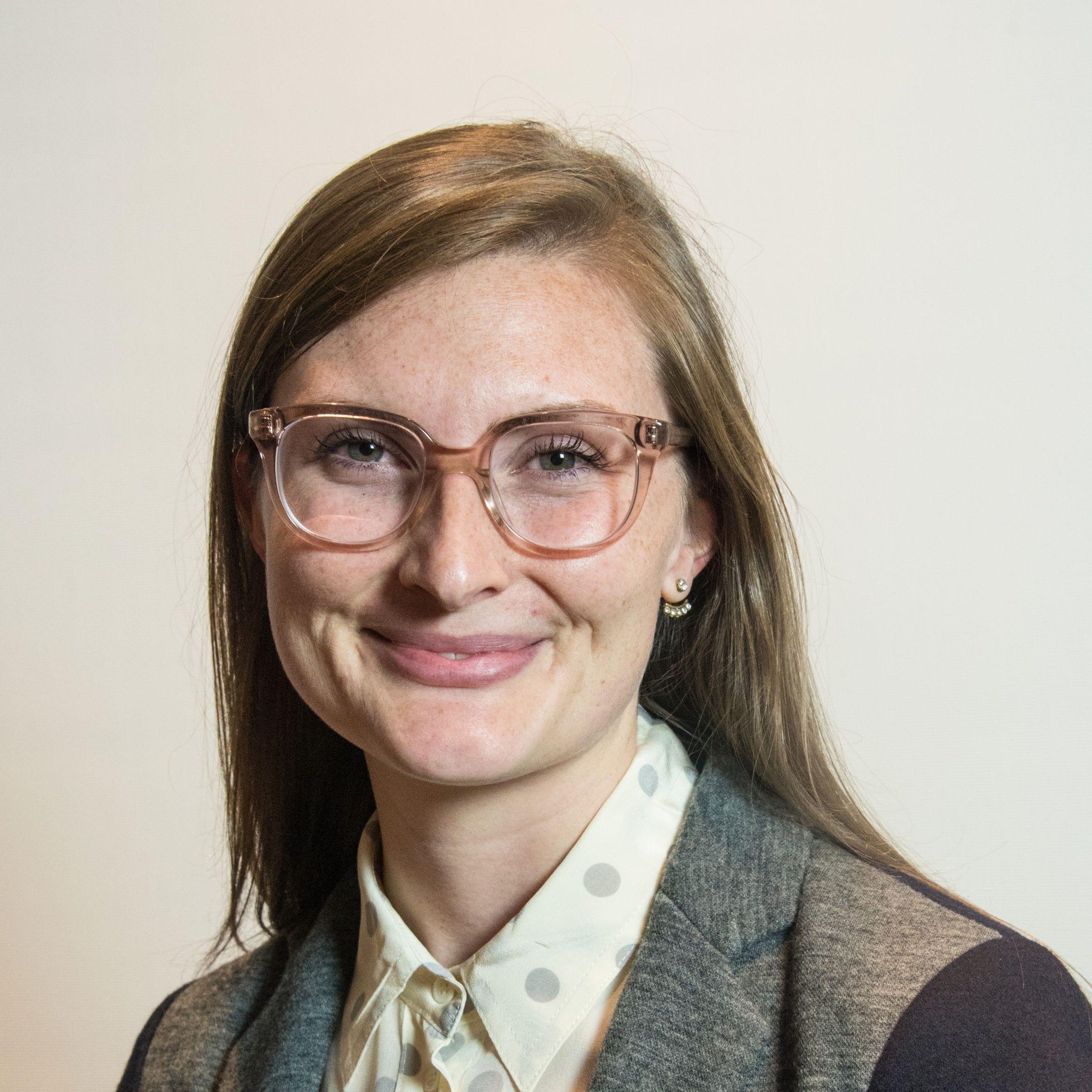 White woman, shoulder-length light brown hair, light brown tinted eyeglasses, polka dot blouse, gray and black blazer