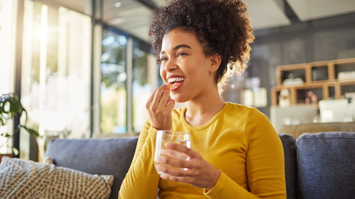 Young adult wearing a sweater and taking a vitamin with a glass of water while seating on the couch.