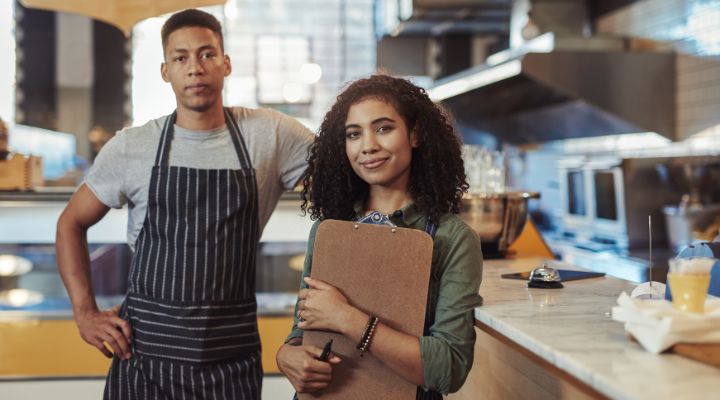 Un hombre con un delantal y una mujer sosteniendo un portapapeles en la cocina de un restaurante.