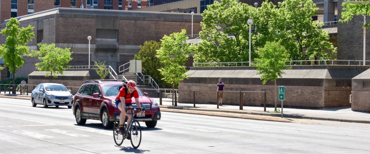 Cars and a biker on a multi-lane road. There is also a person walking on the sidewalk.