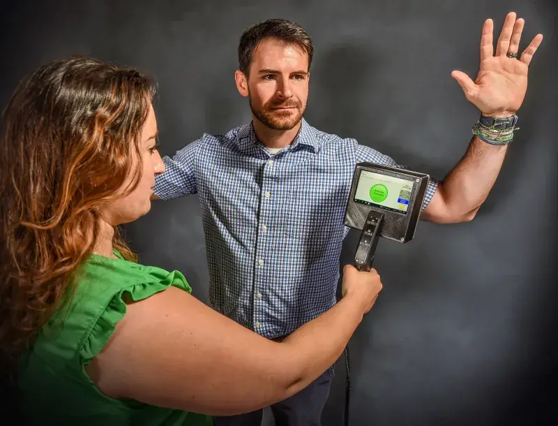 Woman holds up handheld screening wand device near man who stands with arms raised at his sides.