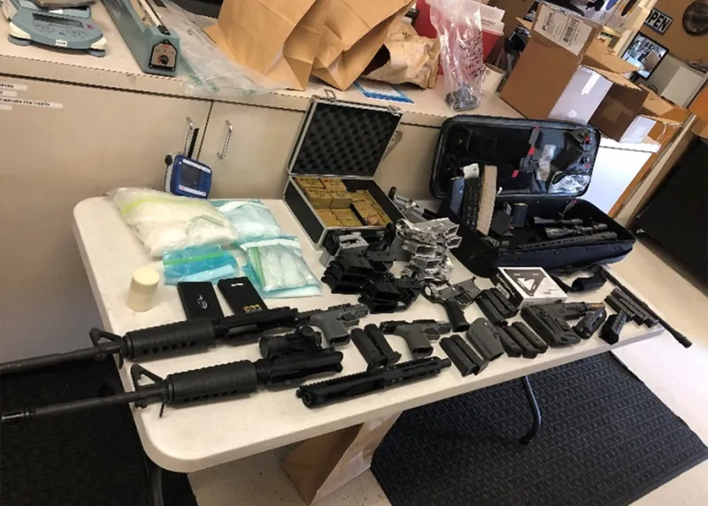 Multiple AR-style firearms, parts, pistols, magazines, unfinished firearm parts used to create ghost guns, and a cache of Russian made ammunition laid out for display on a table.