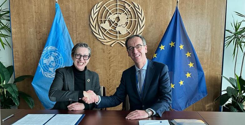 Woman and man shake hand with UN and EU flags in background.