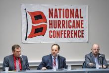 Three men in suits siting behind a table with a banner behind them on the wall. 