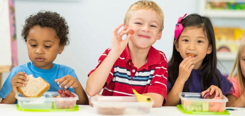 three children at a table smiling and eating healthy food