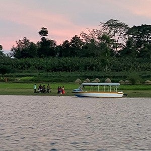 A boat sits docked on the shore of a grassy, forested embankment. A group of people stand on land near the boat.