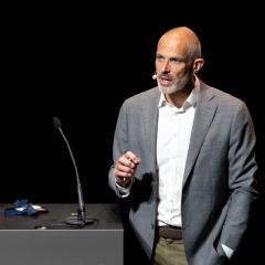 Niklas Blomberg speaking at an event last year. He is wearing a grey jacket and white shirt and is standing next to a black lectern with a microphone on it. Image courtesy of ELIXIR.