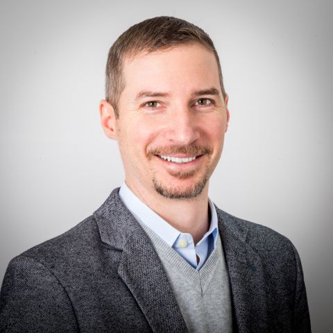 A middle-aged white man with short brown hair and a goatee with beard, both of which are sparkled with white, wearing a dark gray sport coat and light gray sweater vest over a light blue dress shirt, smiling a bad joke he just made.