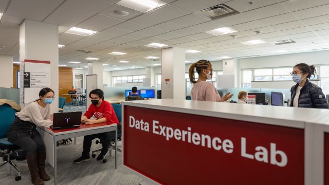 A sign reads "Data Experience Lab" and behind it, people in masks are talking animatedly