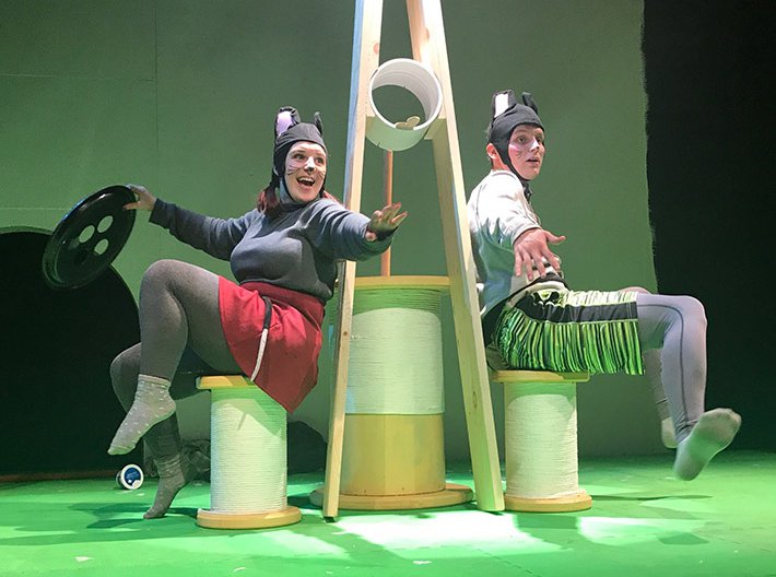 Two actors sitting on stage in between a large clothespin dressed as mice. 