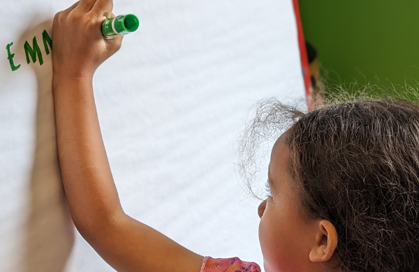 A vhild writing their name on a large white easel with their left hand. 