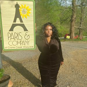 young black woman wearing a black dress with gold hoop earrings and necklace standing by a sign that says Paris and Company