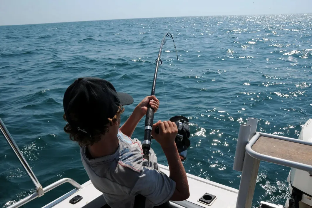 Man wearing a hat pulling in a fish on a fishing rod on a boat 