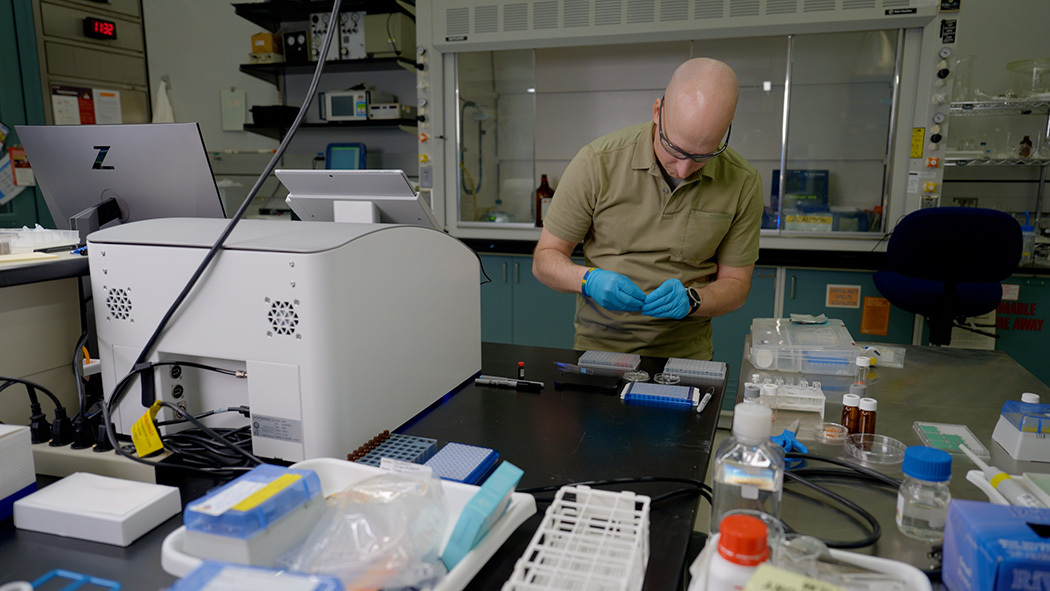 Thomas Forbes stands at a lab table near a white plastic 3D printer, looking down at materials he is preparing. 