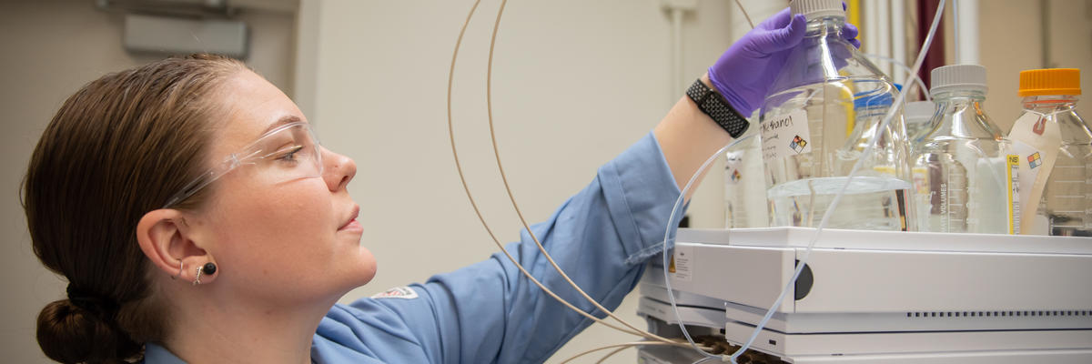 Alix Rodowa wears safety glasses and gloves in the lab as she reaches for a container of clear liquid on top of a piece of scientific equipment. 