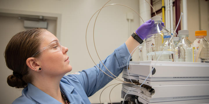 Alix Rodowa wears safety glasses and gloves in the lab as she reaches for a container of clear liquid on top of a piece of scientific equipment. 