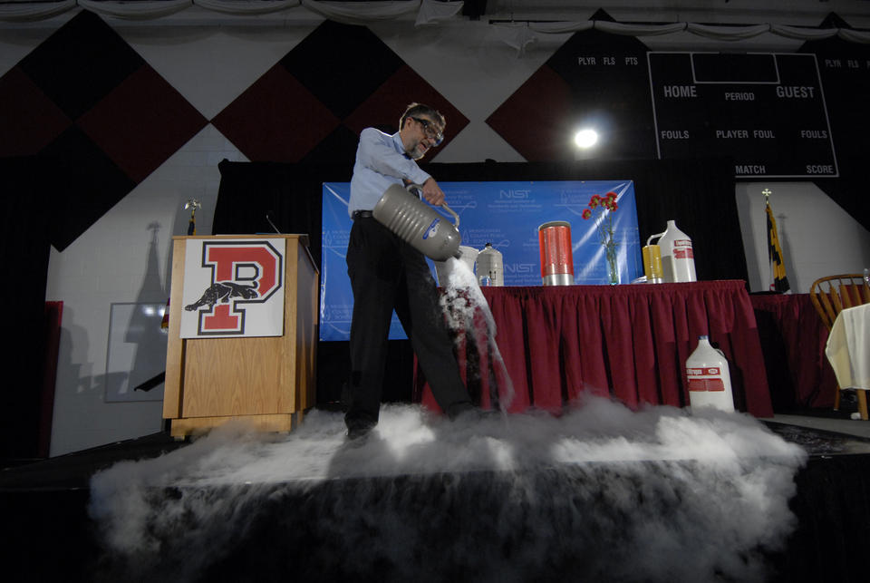 A man in safety glasses pours a foggy-looking liquid out of a metal container as part of a demonstration