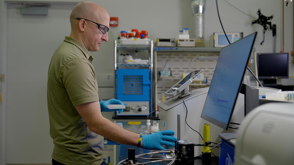 Thomas Forbes wears safety glasses as he looks at the screen attached to a piece of equipment in the lab. 