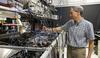 Tasshi Dennis stands in his lab and reaches toward a countertop full of equipment with lots of wires and circuitry.