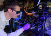 Stephen Eckel wears dark safety glasses as he peers into the complex CAVS apparatus in a darkened lab. 