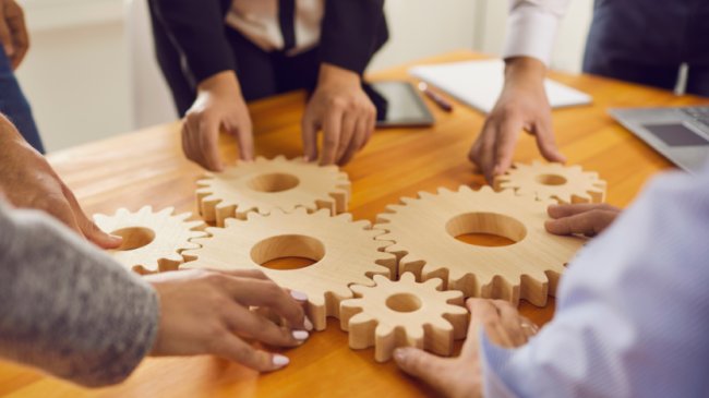 Several pairs of hands fitting wooden gears of various sizes together on a meeting room table. Courtesy of Canva.com