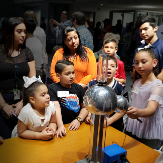 Young children gather around a Van de Graaff machine, a metal structure with a large metal sphere on top, with a metal spherical wand attached. The children watch the machine with faces of awe as they see electricity passing between the machine and the wand.