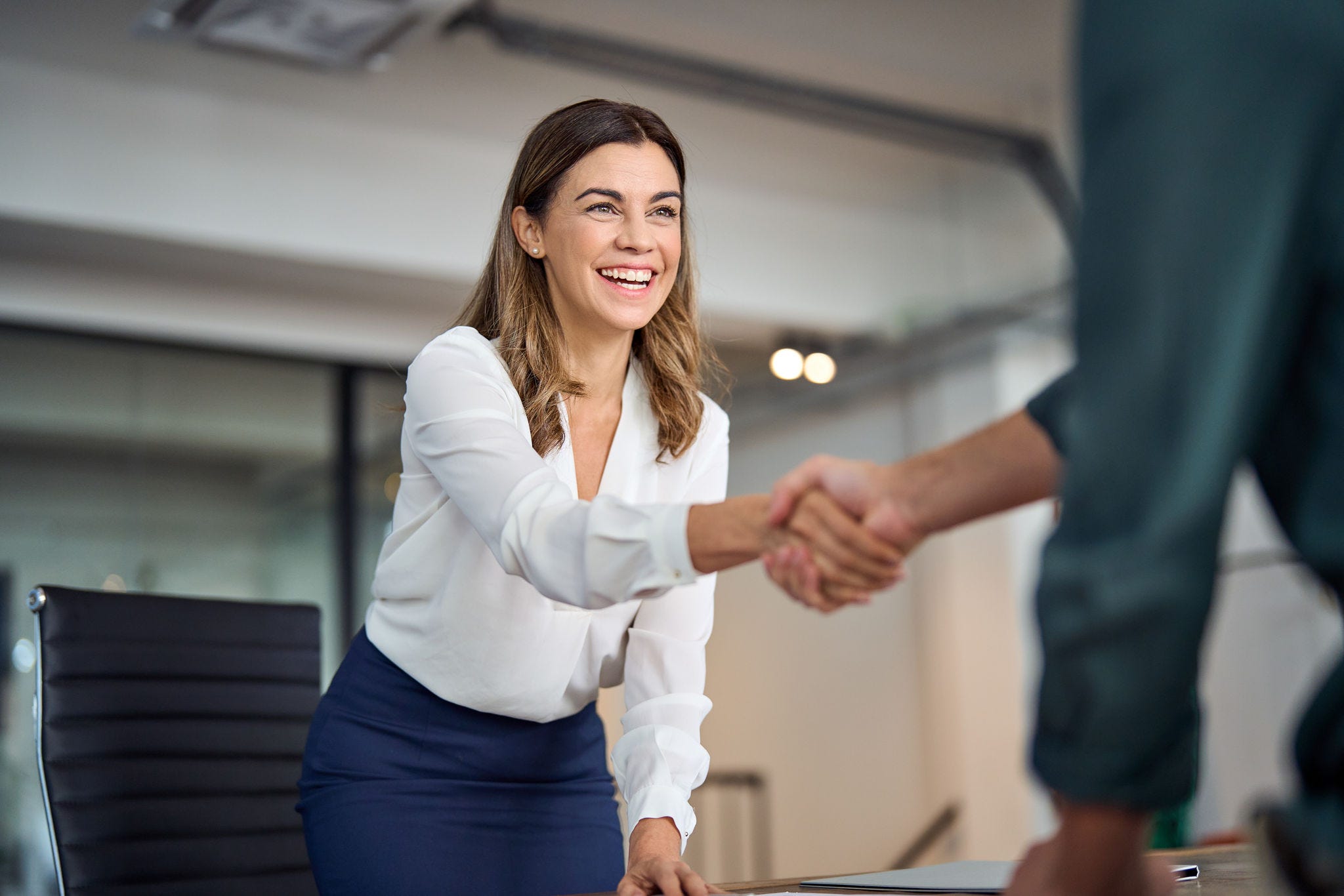 A photograph of a young woman in business dress leaning across a table to shake hands with a colleague, smiling