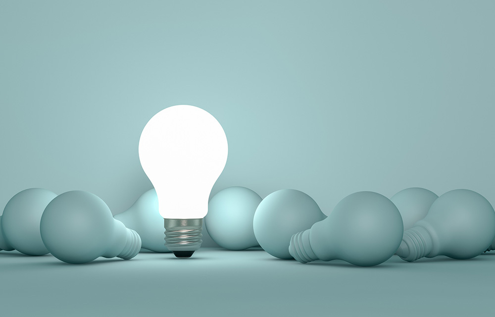 A lit light bulb standing out among a pile of unlit light bulbs on a teal background