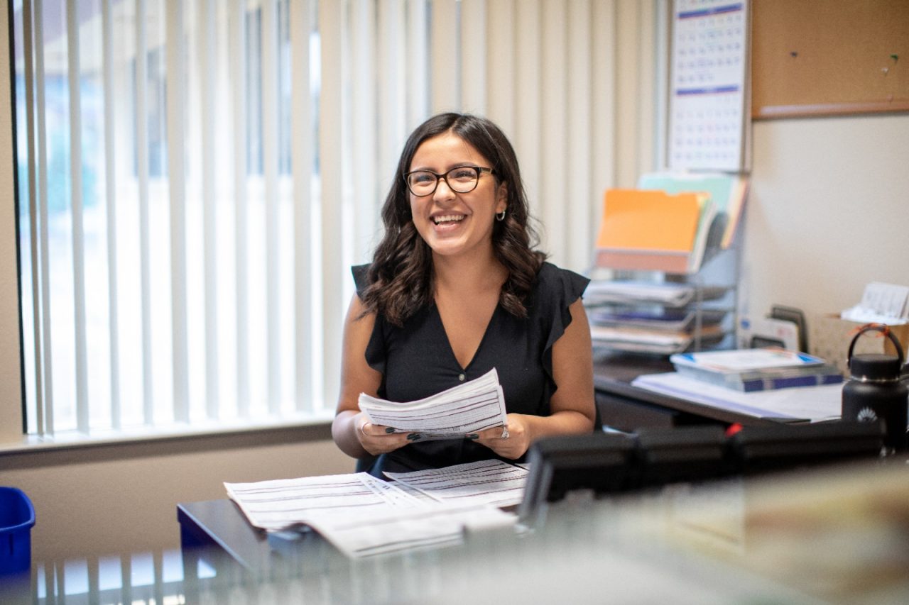 A happpy, young woman sitting at a desk holding paperwork.