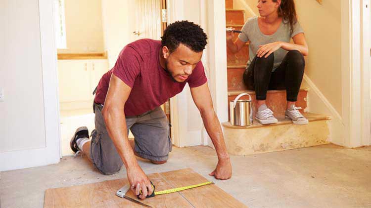 A man measures a board on the floor while a woman paints a wall to help complete their DIY home renovation.