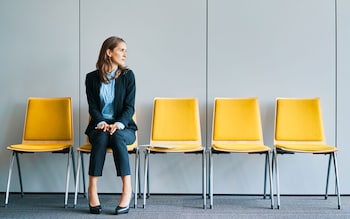 Five yellow chairs are lined up, a woman in a suit sits on the second chair looking to her left