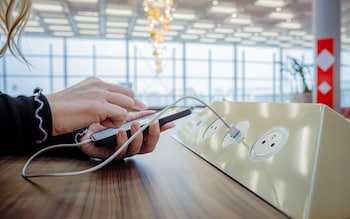 Close-up of a woman with blond hair, wearing a black pullover, using her smartphone while charging it via a white USB cable on a public charging station