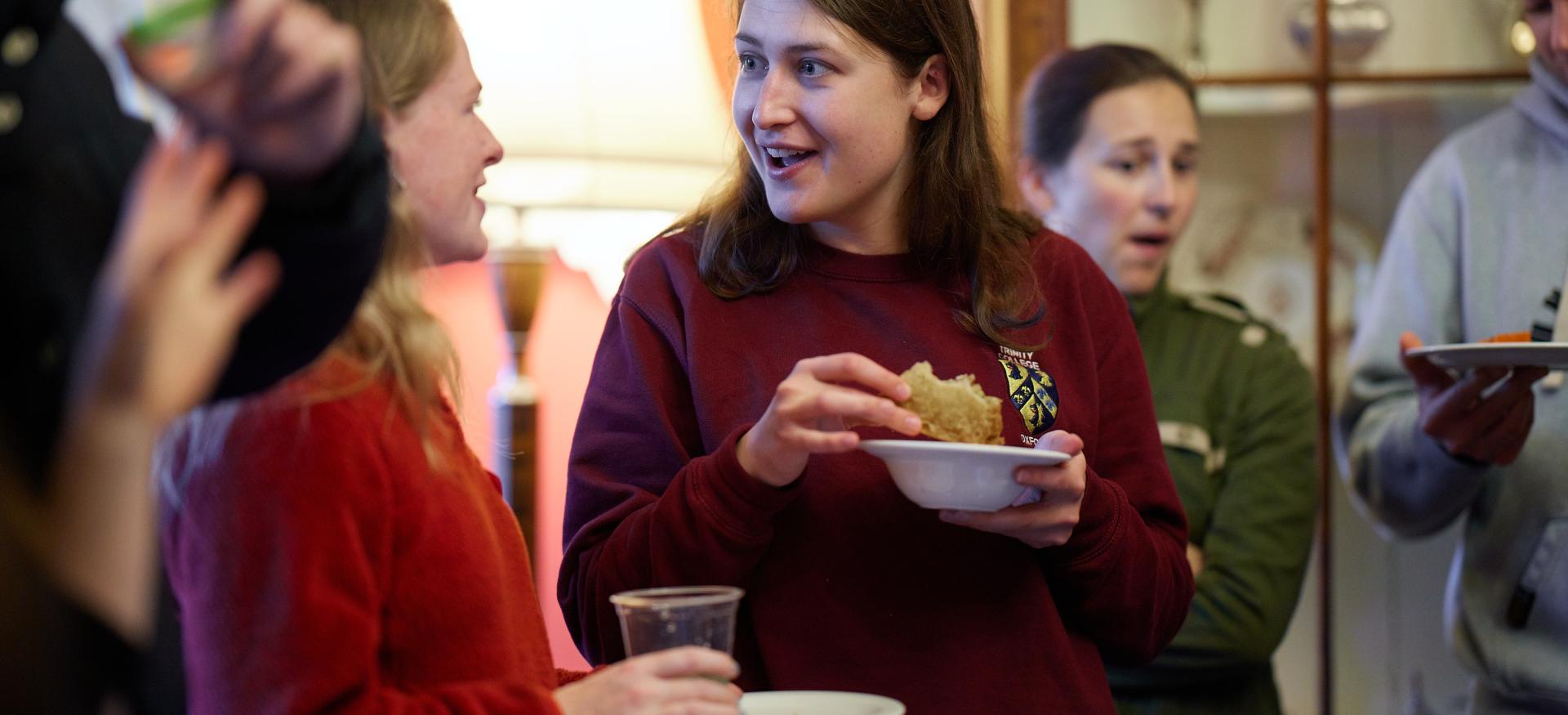 Two female postgraduates stand chatting and eating in the dining room of the Trinity President's Lodgings.