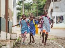 Four girls wearing backpacks run down a dirt and stone road while laughing.