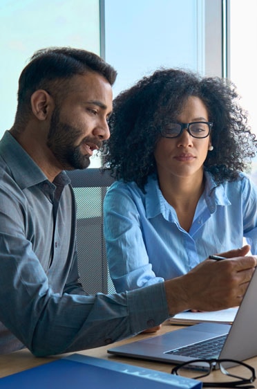 Young man and woman in office setting discussing data on laptop screen