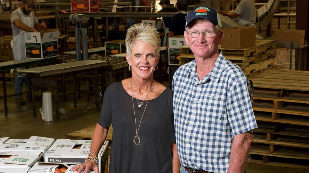 A man and woman stand next to a crate stacked with boxes of sweet potatoes.