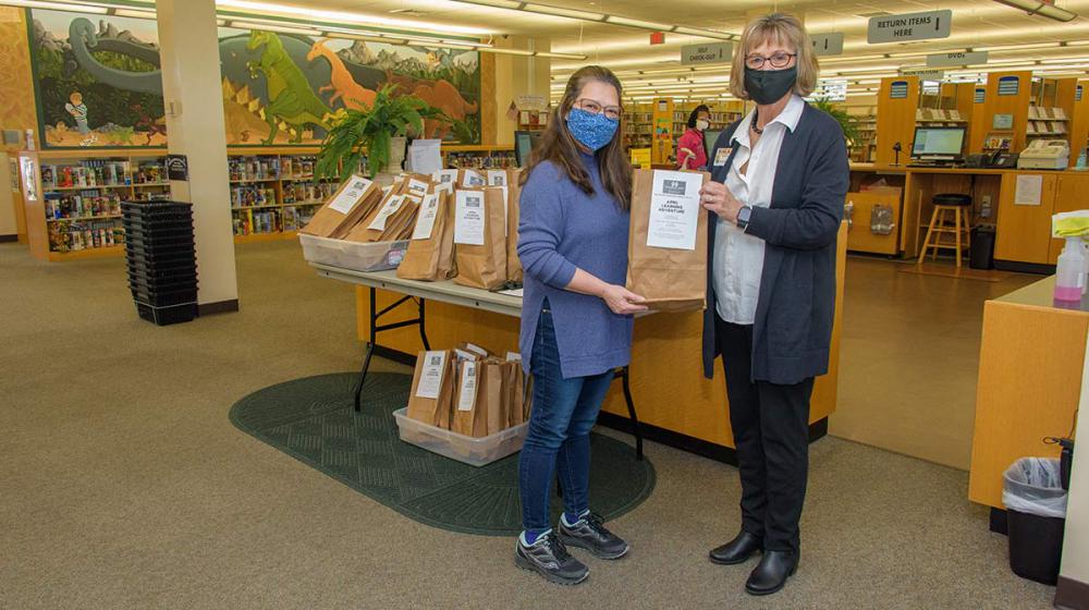 Two masked women with glasses and brown hair holding a brown bag.