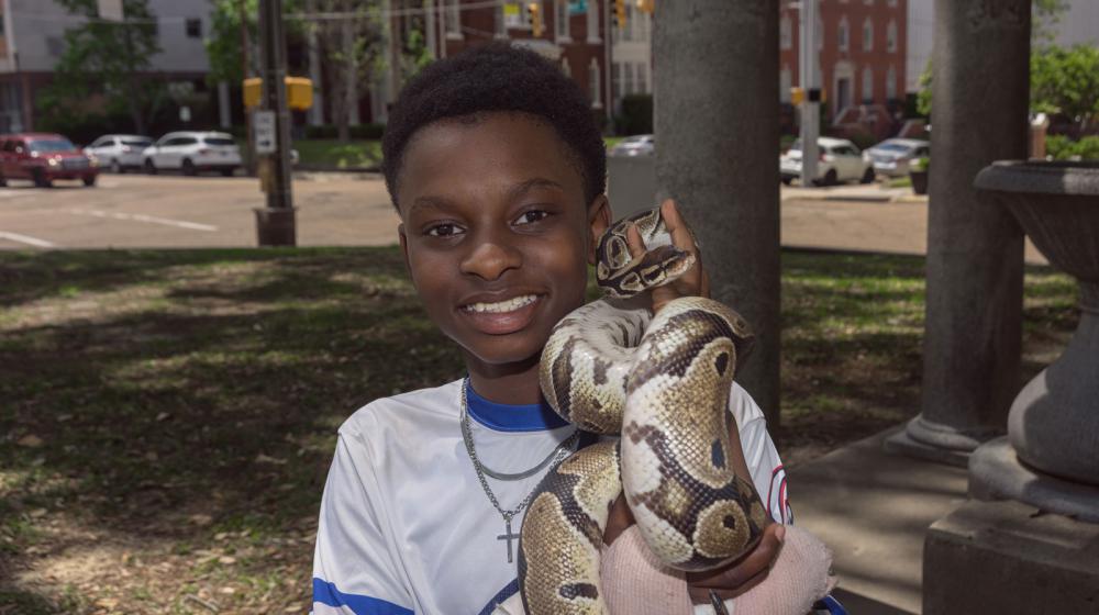 A young smiling Black man holding a snake and standing near a city street.