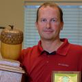 A man wearing a collared red shirt stands holding a large wooden trophy with a large wooden acorn on top in one hand and a plaque in the other.