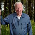 A white-haired male wearing a denim shirt and jeans stands beside a tree and smiles.