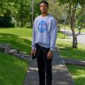 A young boy wearing a NASA sweatshirt stands on a sidewalk holding a camera by his side.