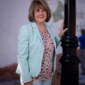 A woman wearing a light blue blazer and pink shirt stands smiling with her hand on a light pole.