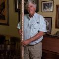 A man holding a large wooden measuring stick stands in front of a wall with several framed photos.