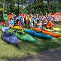 A large group photo of several young children and adults wearing kayaking gear and several kayaks of different colors in the grass in front of them. 