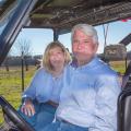 A woman and man seated in a side-by-side with a cattle field stretching behind them.