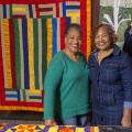 Two Black women smiling and standing in front of a hanging multicolored quilt.