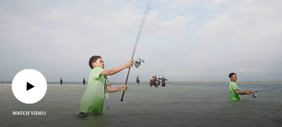 A child casts his fishing reel as he stands hip-deep in the ocean with other kids fishing around him