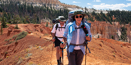 Two people hike along a red-dirt trail at the top of Bryce Canyon National Park while wearing backpacks and using hiking poles