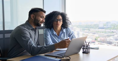 A man and woman look at a laptop computer screen in an office setting.