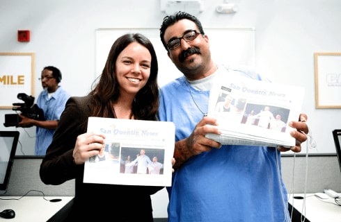Slack volunteer Kathryn Hymes and a person in an incarcerated setting stand smiling alongside each other while both holding up a copy of the San Quentin News.